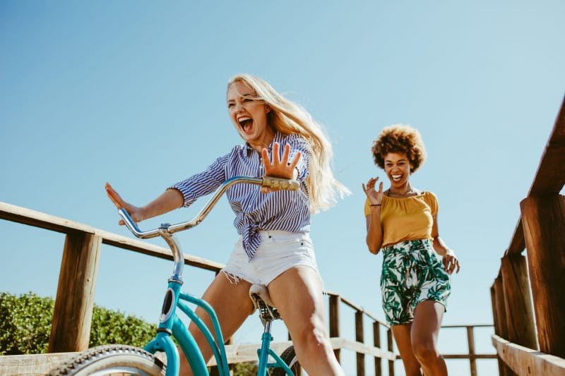 Excited girl cycling on a boardwalk with her friends running. Two woman friends enjoying themselves on the vacation.